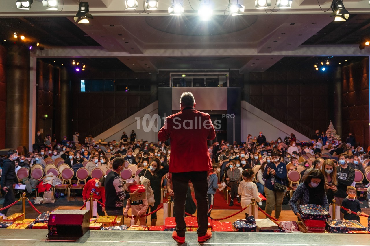 Un homme en veste rouge s'adresse à un public assis dans une grande salle éclairée, avec des enfants au premier plan.
