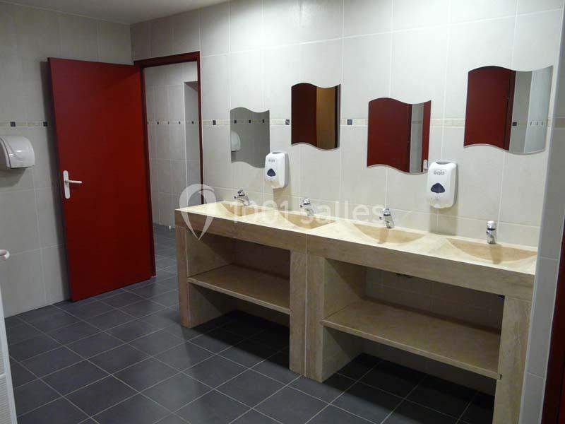 Lavabos alignés avec miroirs dans une salle de bain carrelée, porte rouge ouverte en arrière-plan.