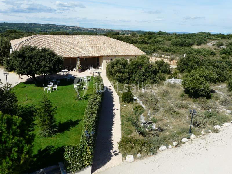 Bâtiment avec toit en tuiles entouré d'un jardin verdoyant, chemin pavé et paysage naturel en arrière-plan.