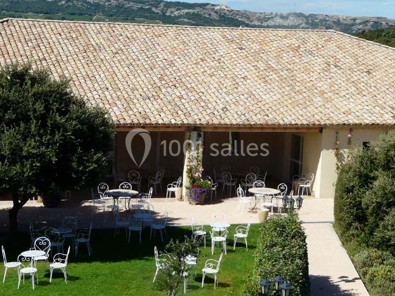 Terrasse avec tables et chaises blanches en fer forgé, entourée de verdure, devant un bâtiment au toit en tuiles.