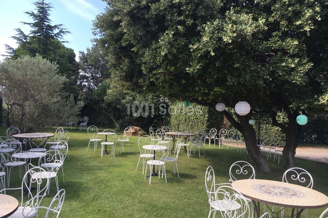 Chaises et tables en fer forgé disposées sur une pelouse, entourées d'arbres et de lanternes suspendues.
