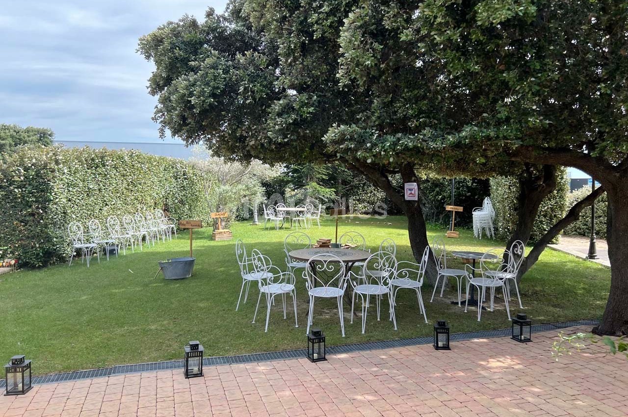 Jardin aménagé avec des tables et chaises blanches en fer forgé, sous des arbres et entouré de haies.
