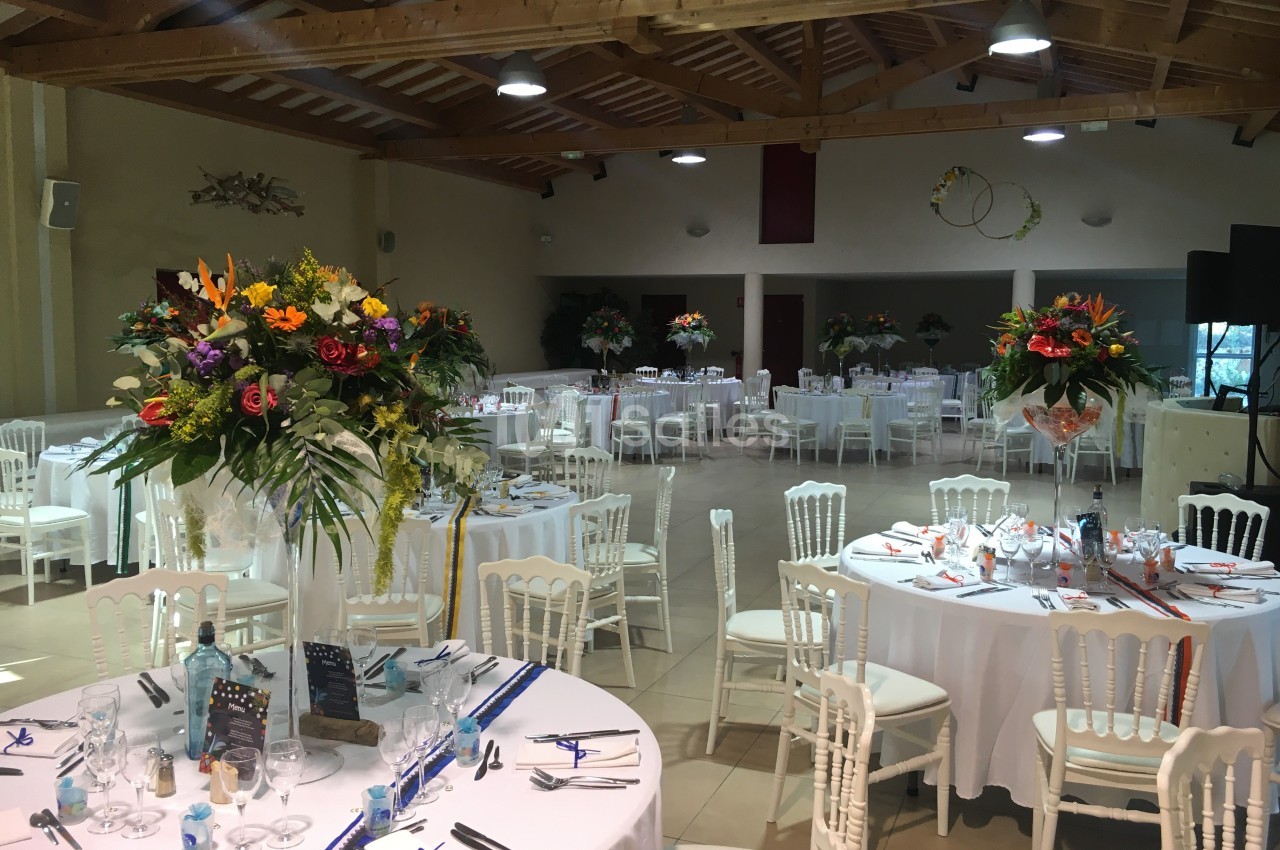 Salle de réception décorée avec des tables rondes dressées, ornées de bouquets de fleurs colorées.