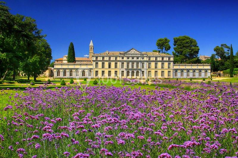 Façade d'un grand bâtiment historique entouré de jardins fleuris et d'arbres sous un ciel bleu.