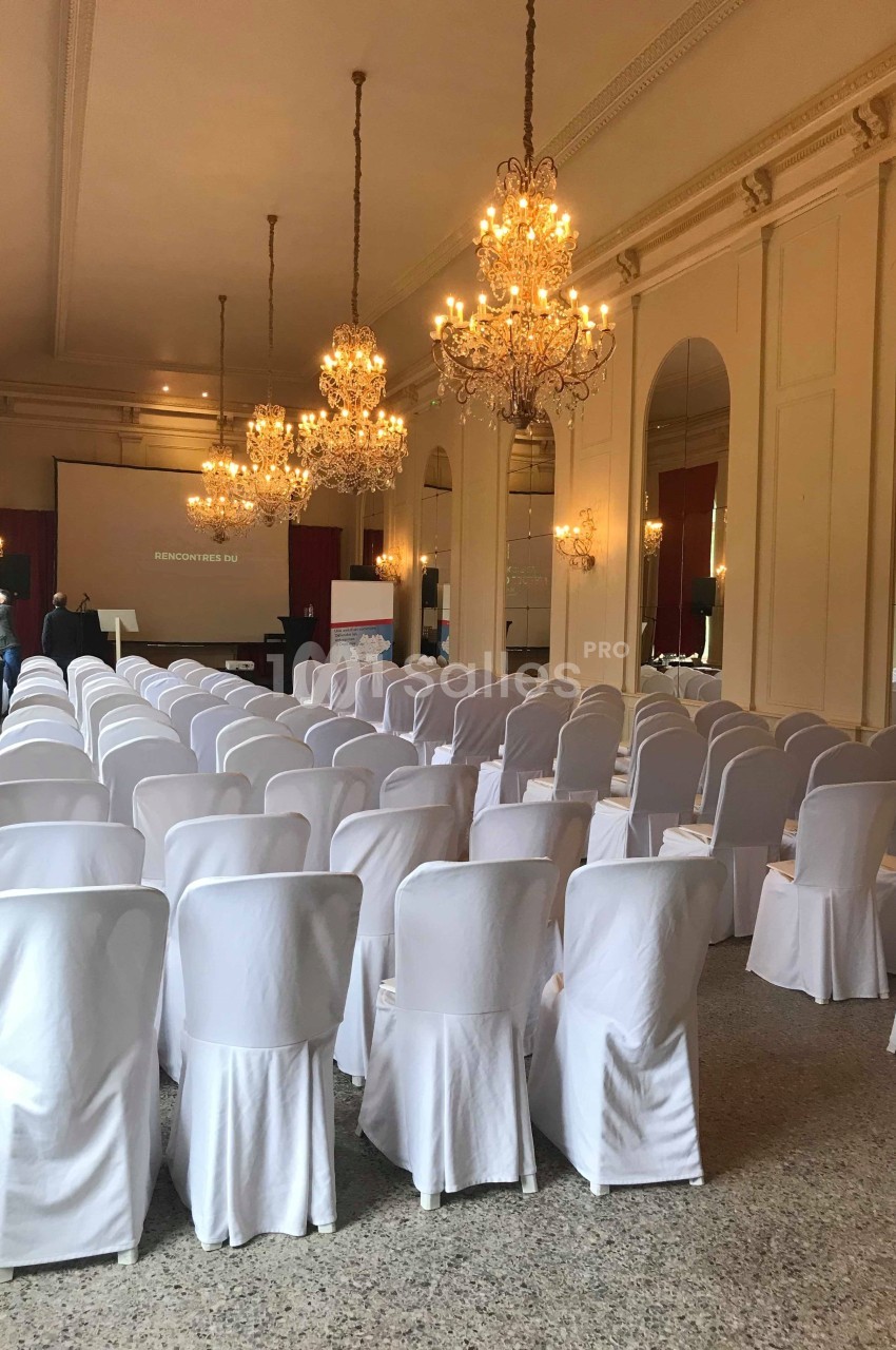 Salle de conférence élégante avec rangées de chaises blanches, lustres suspendus et écran de projection à l'arrière.