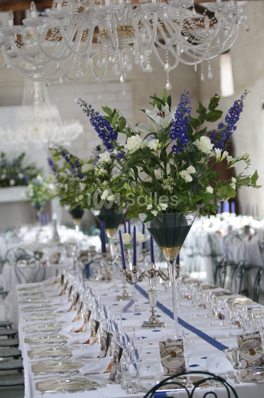 Grande table de réception élégamment dressée avec chandeliers, verres, et compositions florales bleues et blanches.