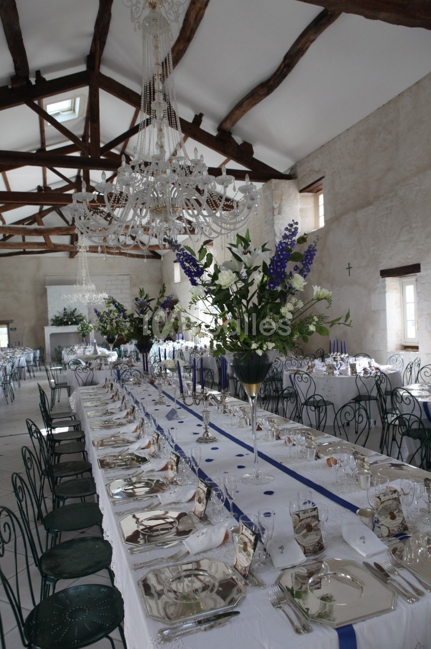 Salle de réception décorée avec une longue table dressée, ornée de fleurs blanches et violettes sous des lustres en cristal.