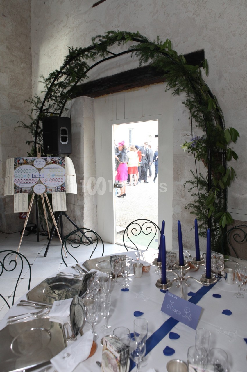 Table dressée avec des chandeliers bleus sous une arche végétale, donnant sur une porte ouverte vers des invités à l…