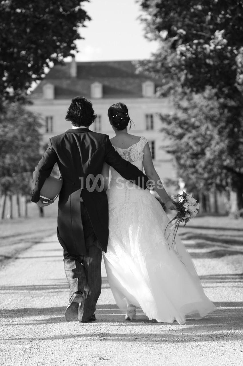 Un couple de mariés marchant sur une allée bordée d'arbres, vu de dos, devant un bâtiment élégant.