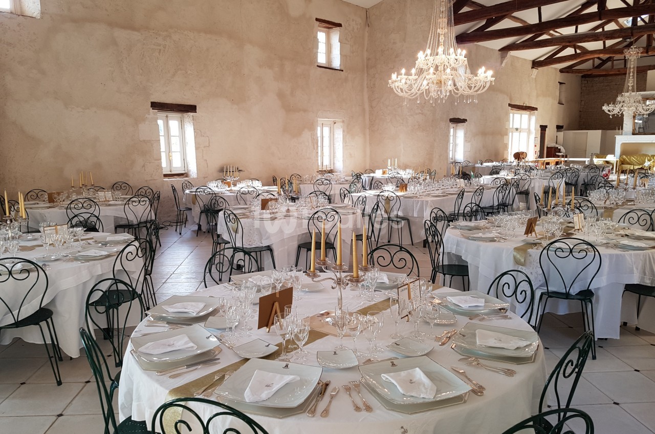 Salle de réception décorée avec des tables rondes dressées, nappes blanches, chandeliers dorés et chaises en fer forgé.