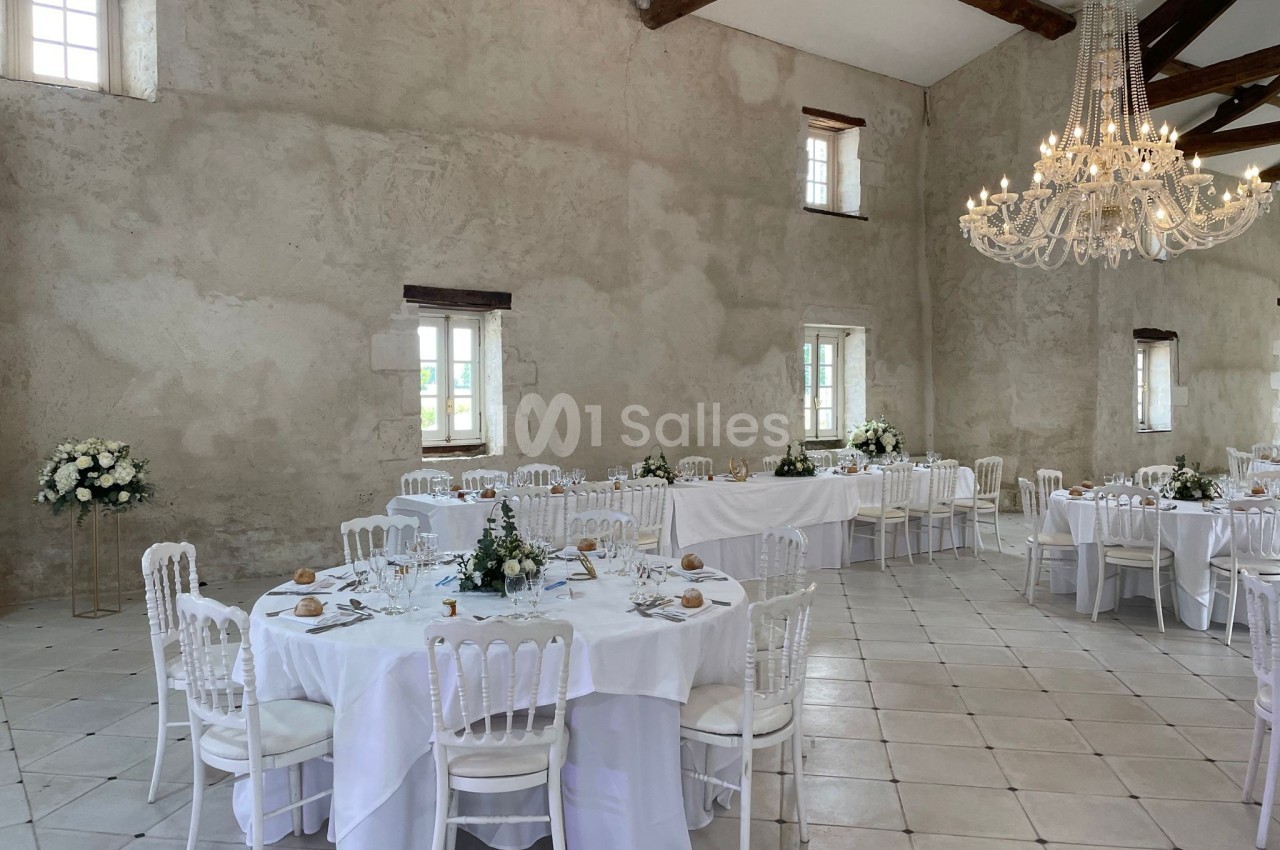 Salle de réception lumineuse avec tables rondes et rectangulaires décorées de nappes blanches et arrangements floraux.
