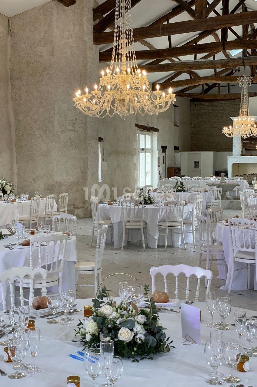 Salle de réception lumineuse avec tables décorées de fleurs blanches, chaises blanches et lustres élégants.