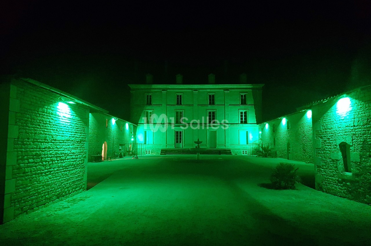 Façade d'un bâtiment éclairé en vert la nuit, entouré de murs en pierre et d'une cour centrale.