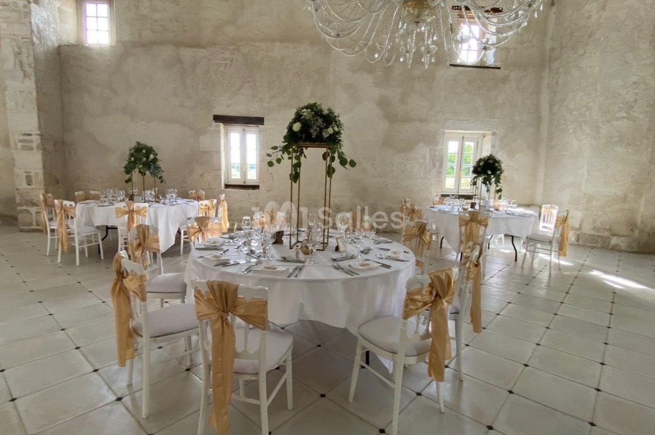 Salle de réception lumineuse avec tables rondes décorées de nappes blanches, chaises ornées de rubans dorés et centre de…