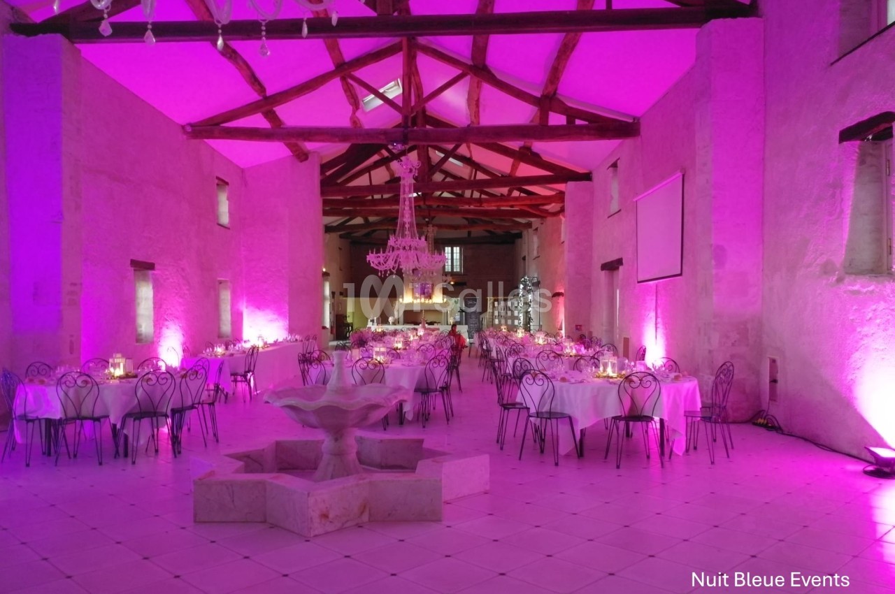 Salle de réception avec tables rondes dressées, éclairage rose, lustre central et fontaine en pierre au premier plan.
