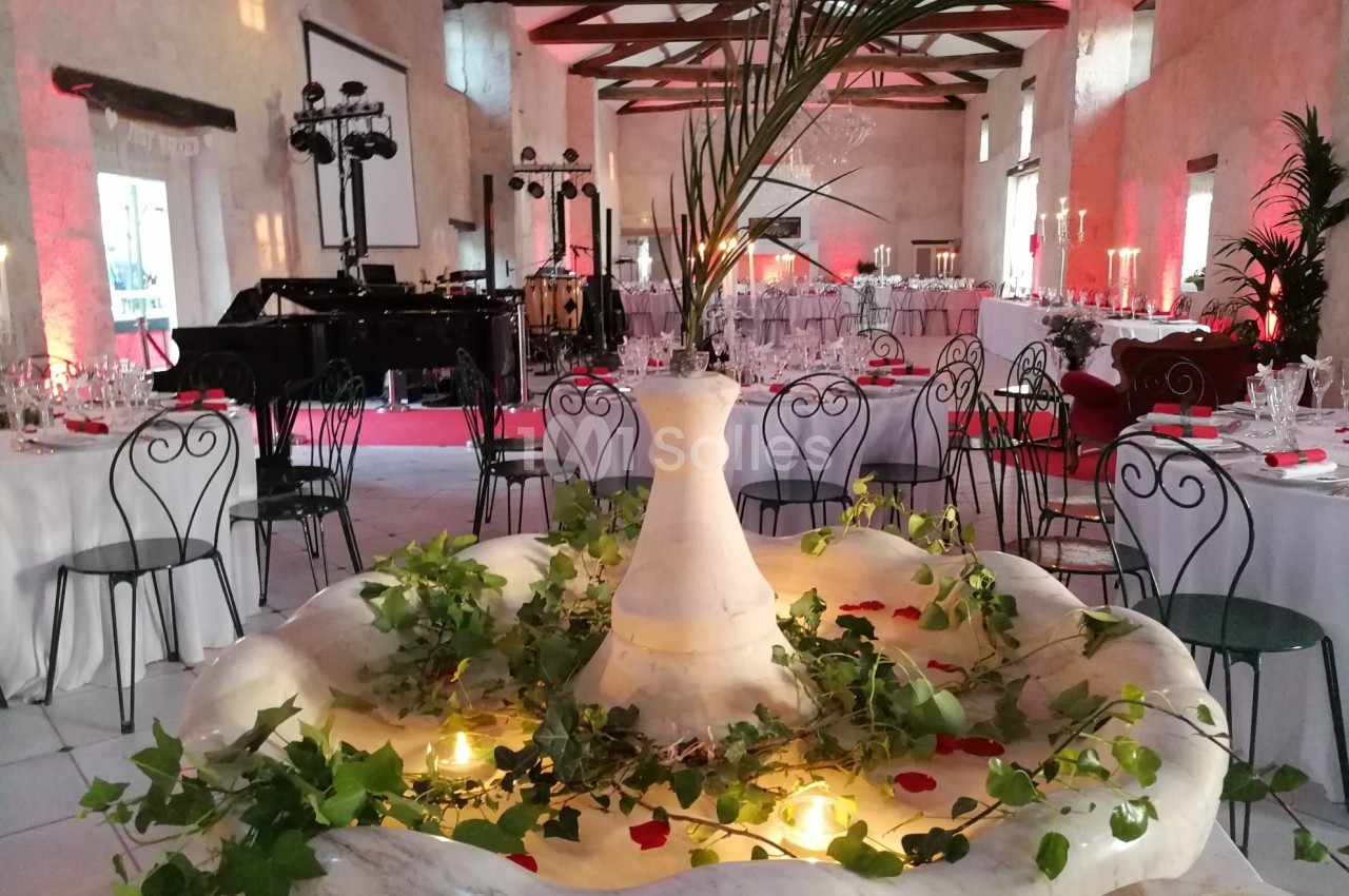 Salle de réception décorée avec des tables dressées, une fontaine ornée de feuillage et de bougies au premier plan.