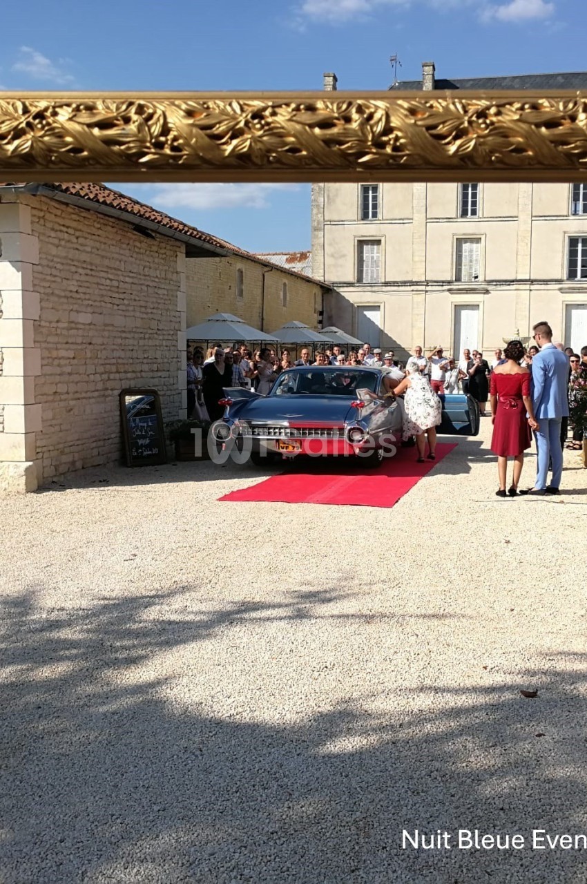 Cadre doré entourant une scène de mariage avec une voiture ancienne, un tapis rouge et des invités devant un bâtiment en…