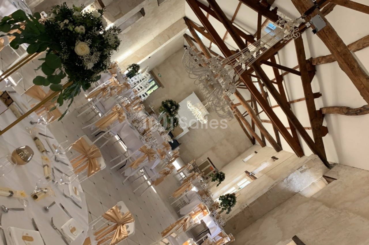 Salle de réception décorée pour un mariage, avec tables dressées, chaises ornées de nœuds dorés et lustres suspendus.