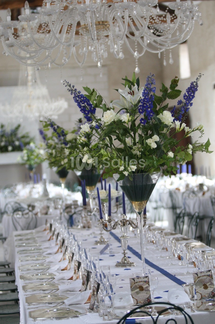 Grande table de réception élégamment dressée avec chandeliers, verres alignés et bouquets de fleurs blanches et bleues.