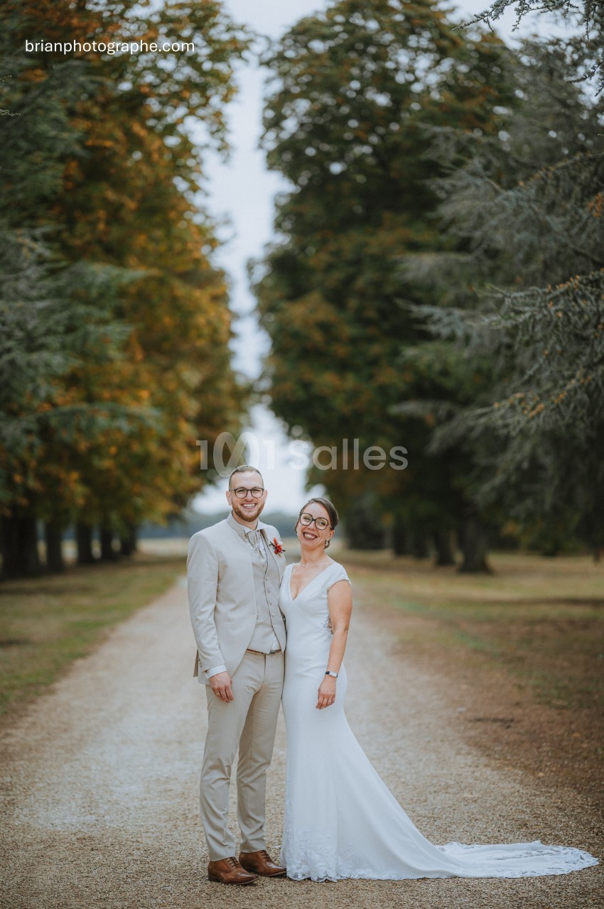 Un couple en tenue de mariage pose sur une allée bordée d'arbres dans un cadre naturel.
