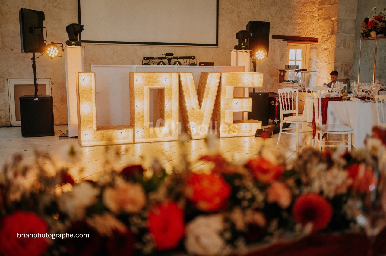 Lettres lumineuses formant le mot ’LOVE’ dans une salle décorée pour un événement, avec des fleurs au premier plan.