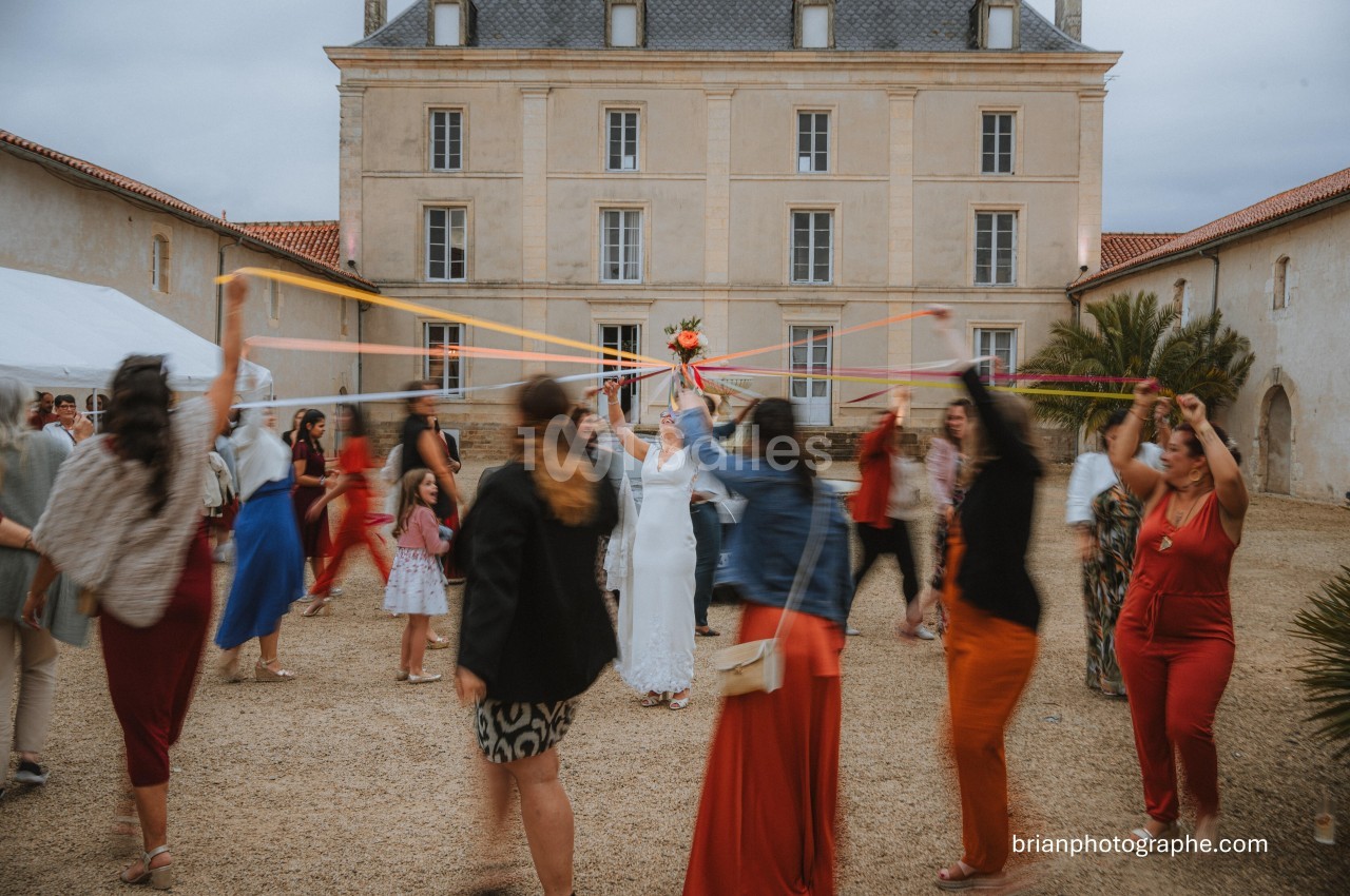 Des invités dansent en cercle autour de la mariée, tenant des rubans colorés, devant un bâtiment ancien.