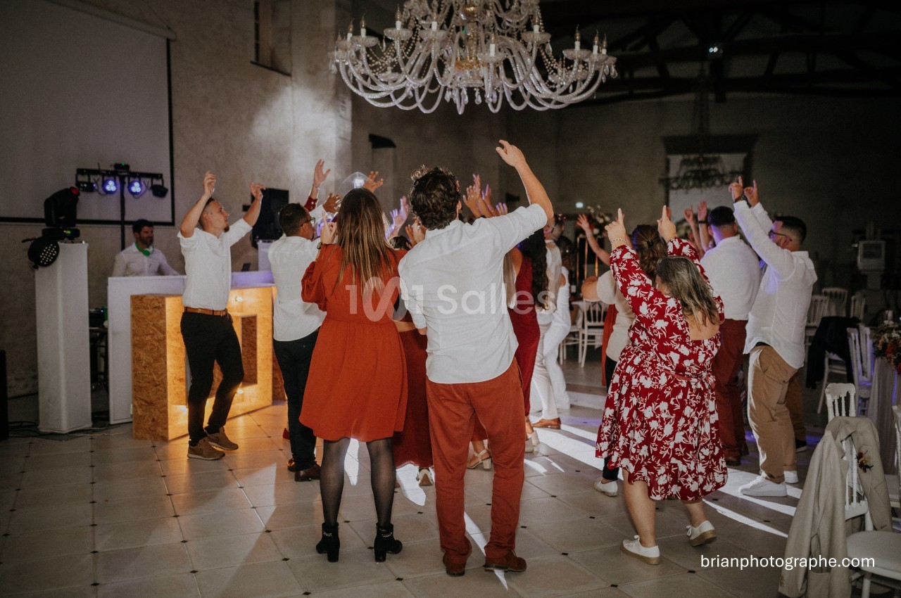 Des invités dansent sous un lustre dans une salle de réception, éclairée par des lumières de fête.