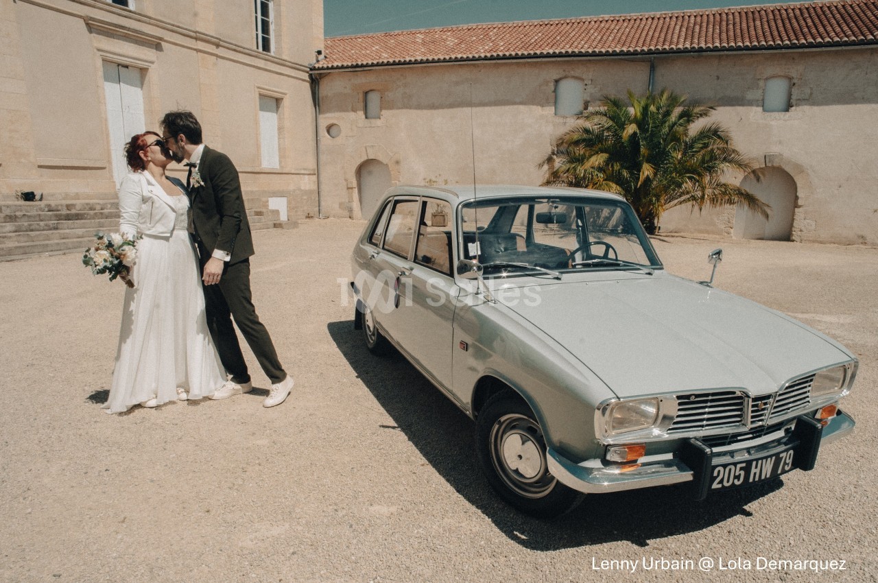 Un couple de mariés s'embrasse devant une voiture ancienne stationnée dans une cour ensoleillée.