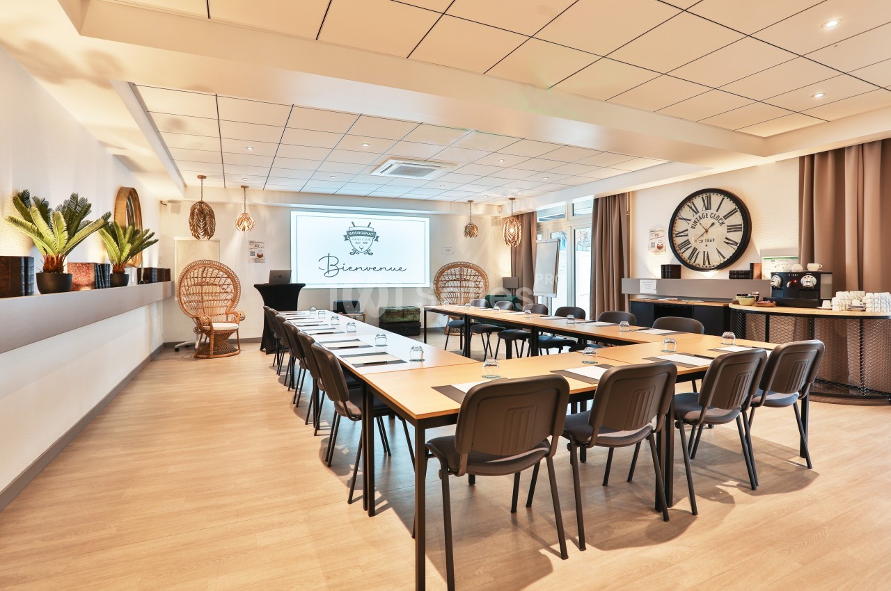 Salle de réunion lumineuse avec tables disposées en U, écran de projection, décorations et coin café.