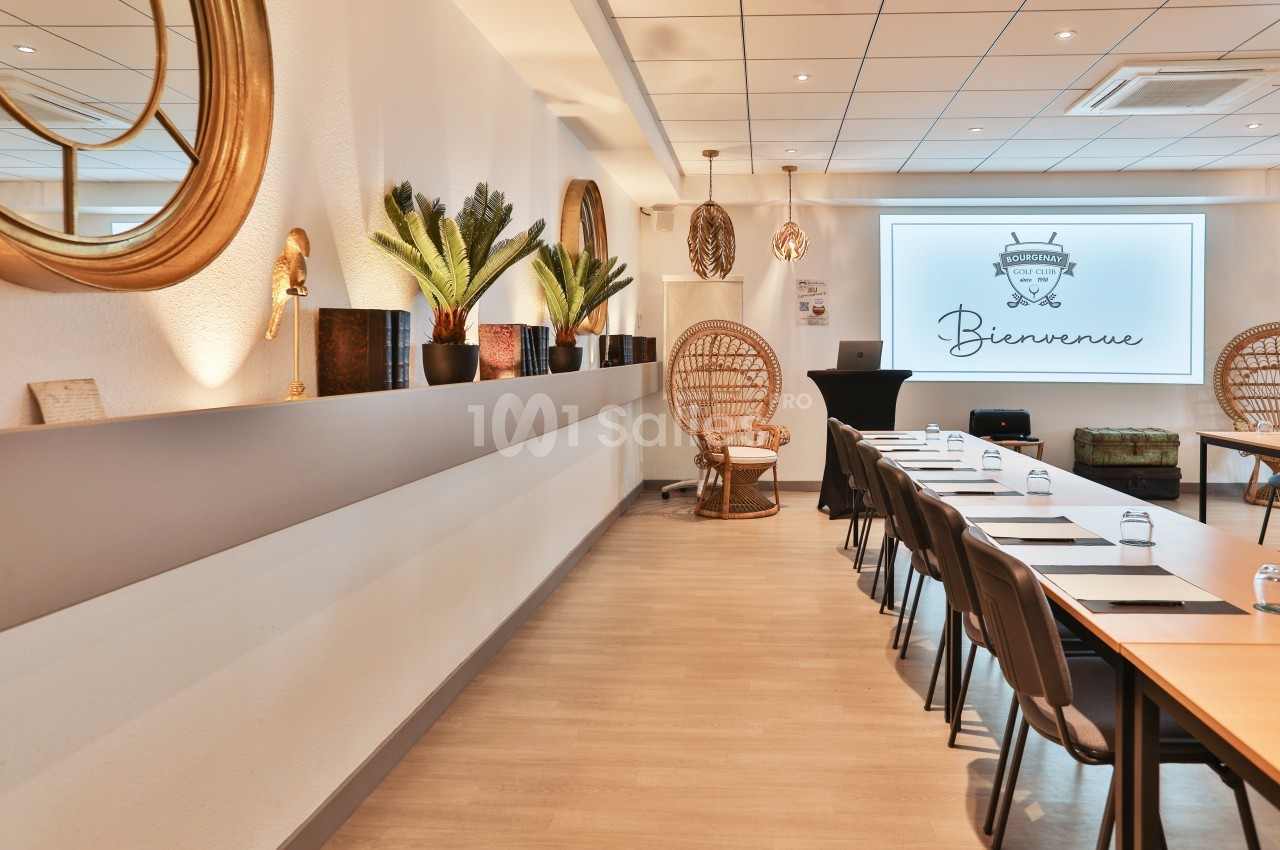 Salle de réunion lumineuse avec tables alignées, chaises noires, plantes décoratives et écran affichant ’Bienvenue’.