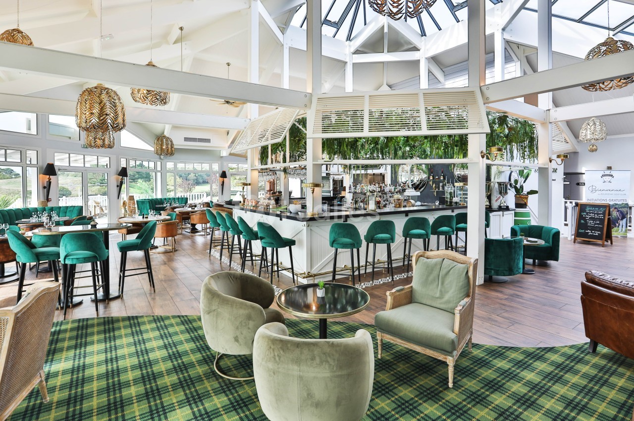 Intérieur lumineux d'un bar-restaurant avec mobilier vert, comptoir central, plantes suspendues et grandes baies vitrées.
