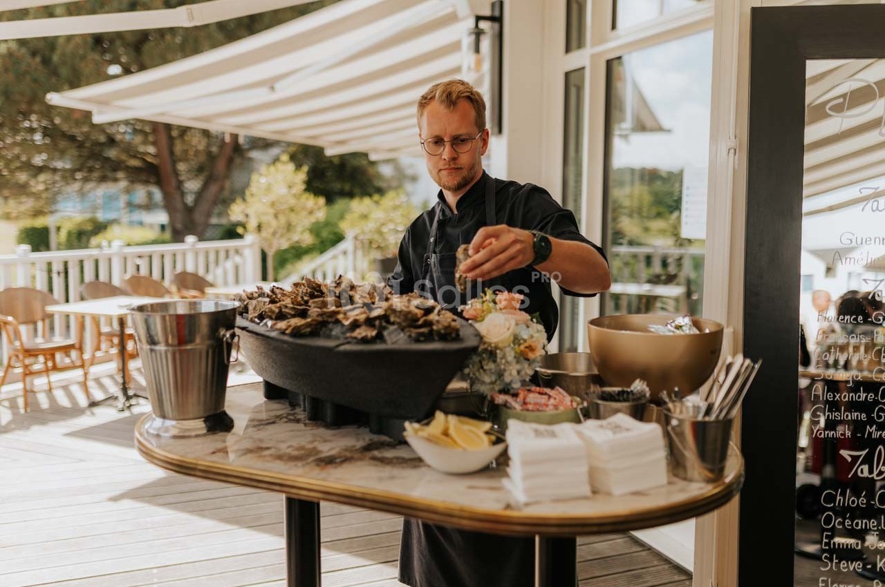 Un homme prépare un plateau d'huîtres sur une terrasse ensoleillée, avec des citrons et des serviettes à disposition.