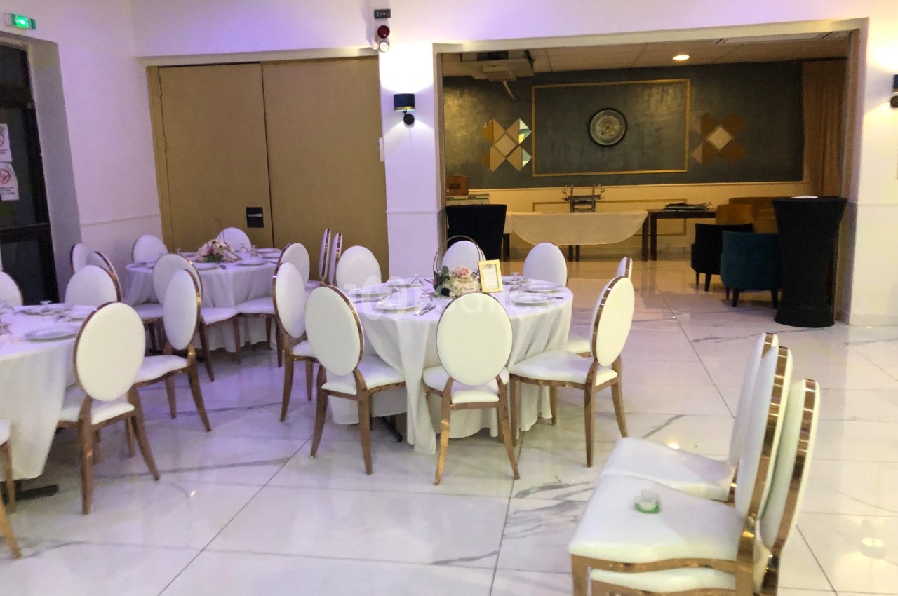 Salle de réception lumineuse avec tables rondes dressées, chaises blanches et décor sobre en arrière-plan.