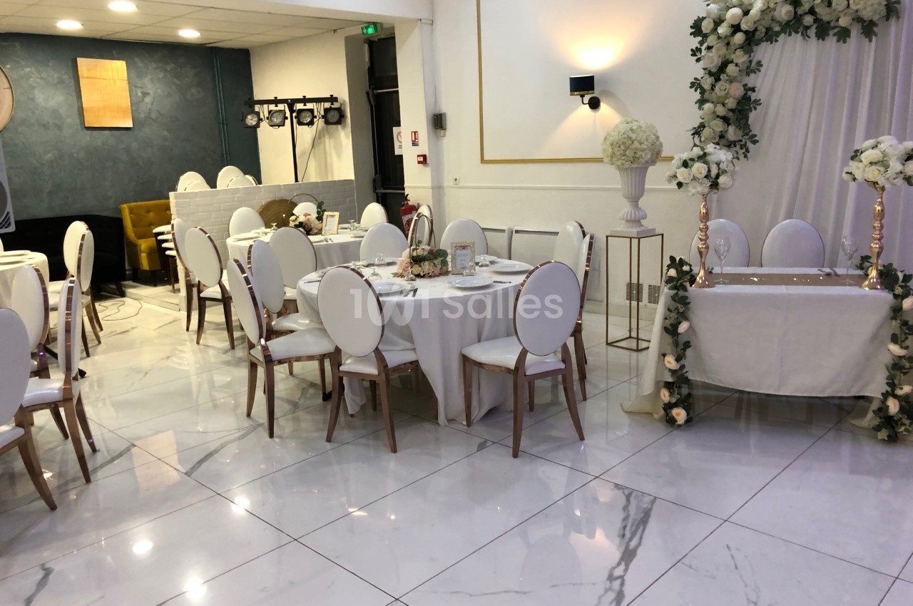 Salle de réception élégante avec tables rondes dressées, chaises blanches et arche florale décorative.