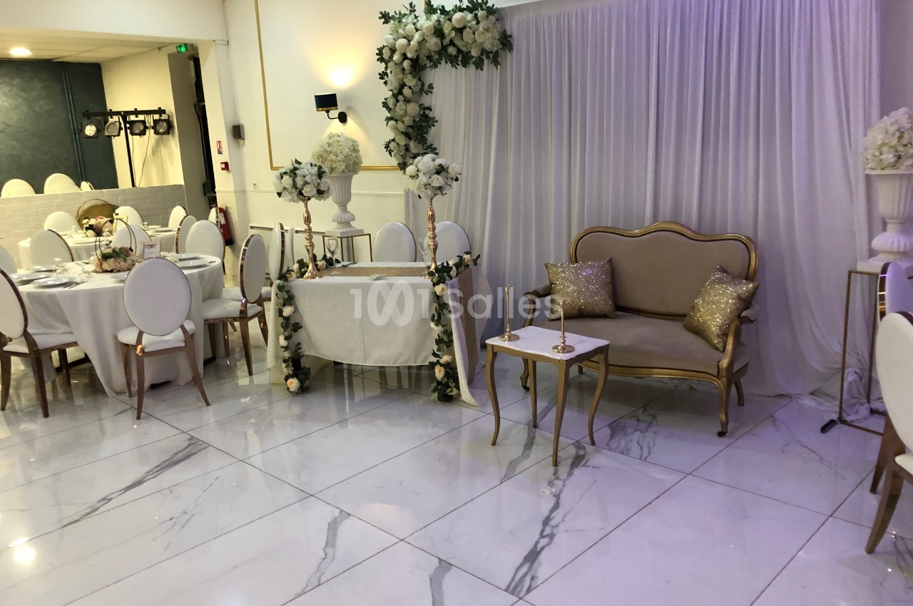Salle de réception décorée avec des tables rondes, un espace d'honneur avec arche florale et un canapé doré.