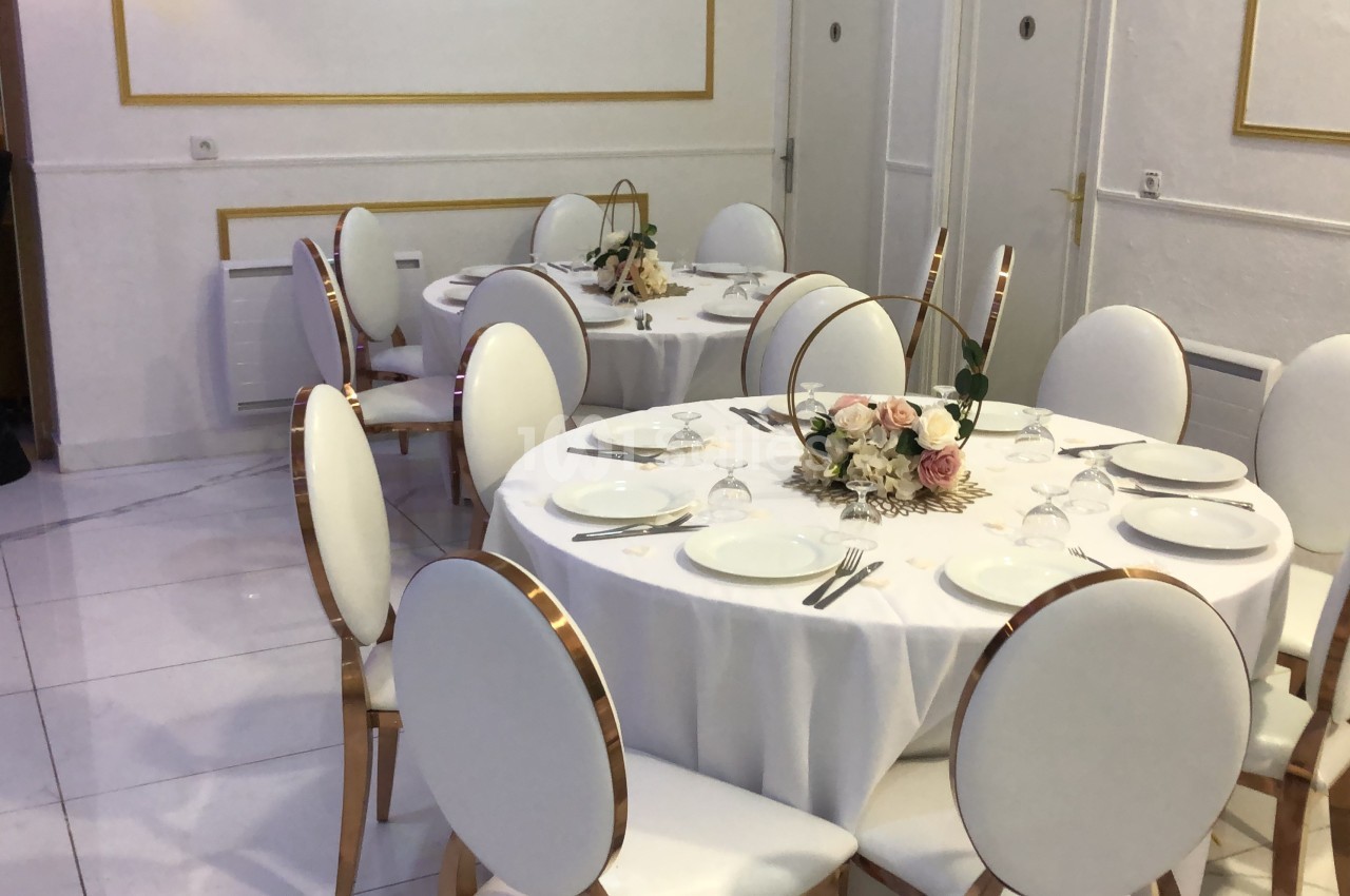 Salle élégante avec tables rondes dressées, nappes blanches, chaises assorties et centres de table floraux.