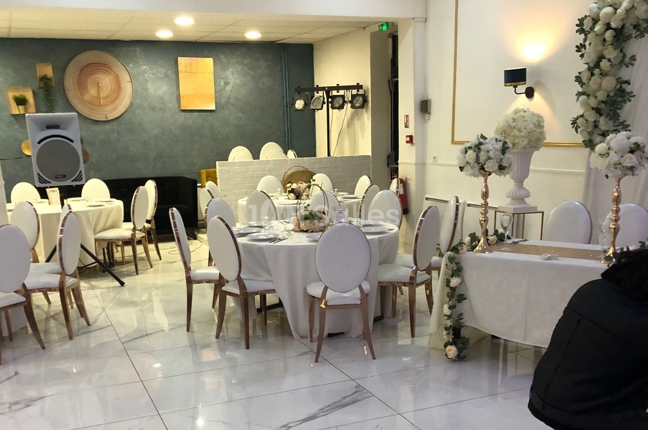 Salle de réception élégante avec tables rondes dressées, chaises blanches et décoration florale raffinée.