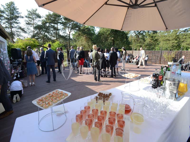 Buffet en extérieur avec boissons et amuse-bouches, des invités discutent sous un grand parasol sur une terrasse ensoleillée.
