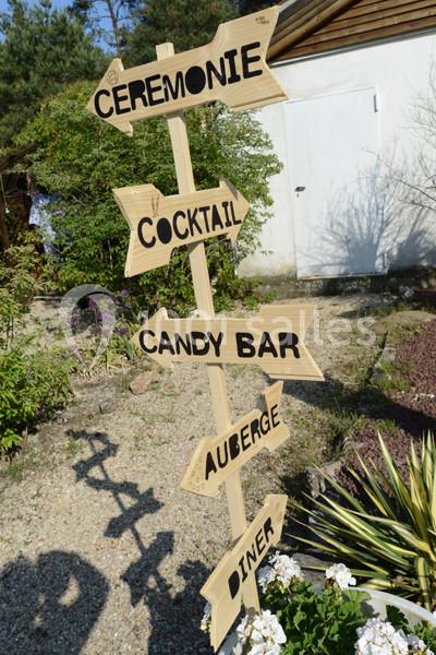 Panneau en bois avec des flèches indiquant ’Cérémonie’, ’Cocktail’, ’Candy Bar’, ’Auberge’ et ’Dîner’, dans un jardin.