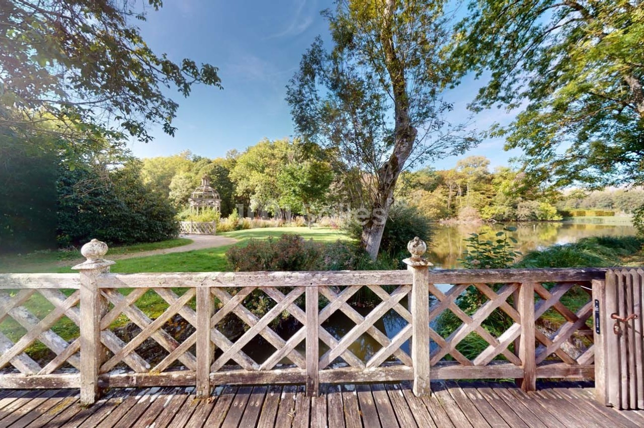 Vue depuis une terrasse en bois avec une balustrade, donnant sur un parc arboré et un étang sous un ciel dégagé.