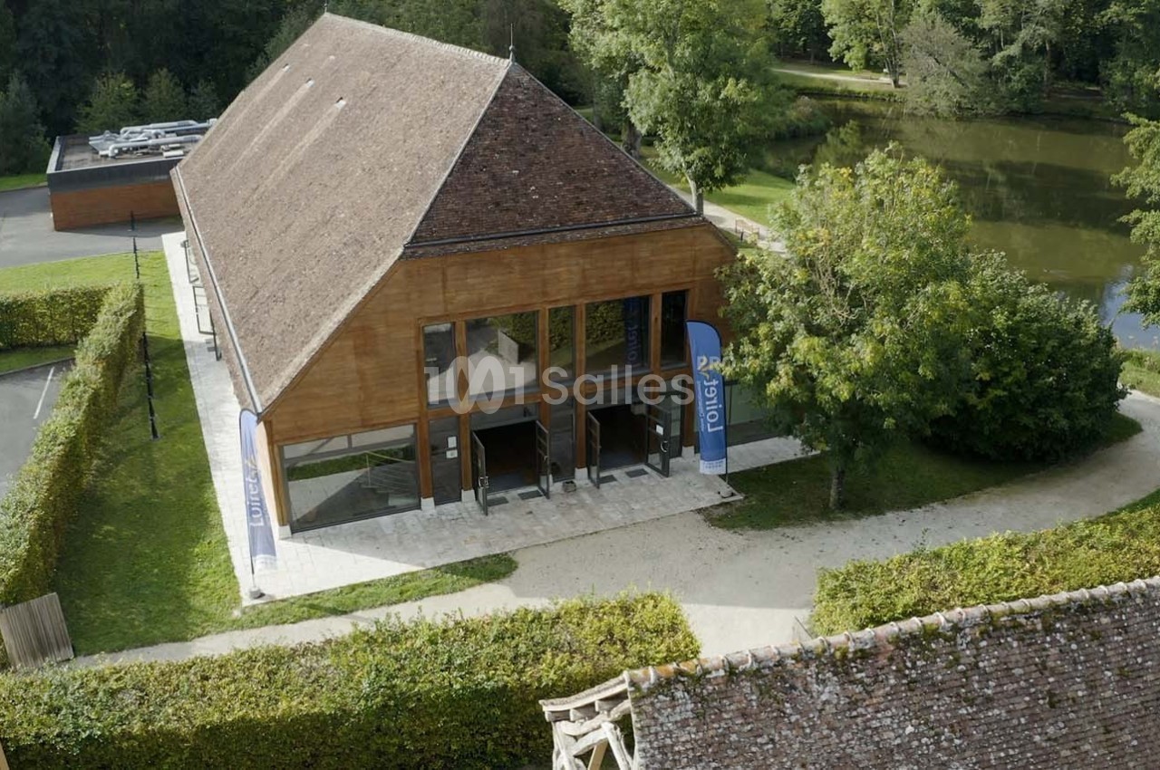 Bâtiment en bois avec de grandes baies vitrées, entouré de verdure, près d'un étang et d'un parking.