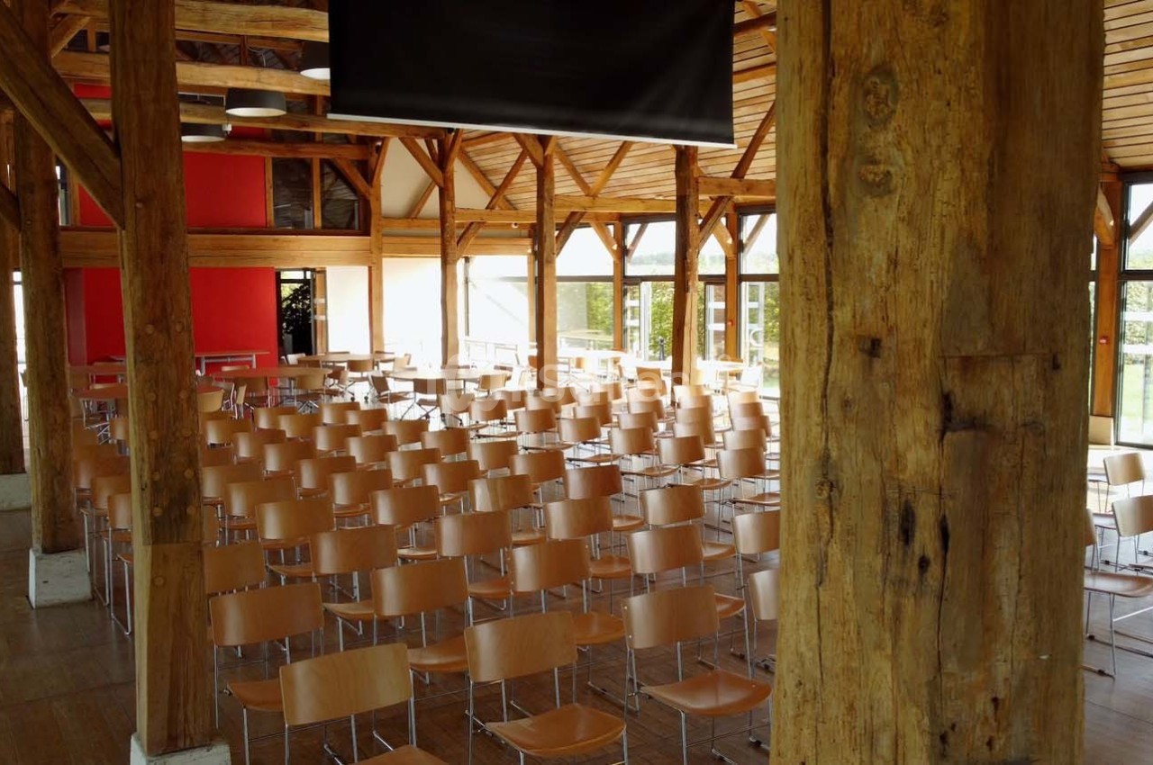 Salle de conférence lumineuse avec des rangées de chaises en bois clair, poutres apparentes et grandes fenêtres.