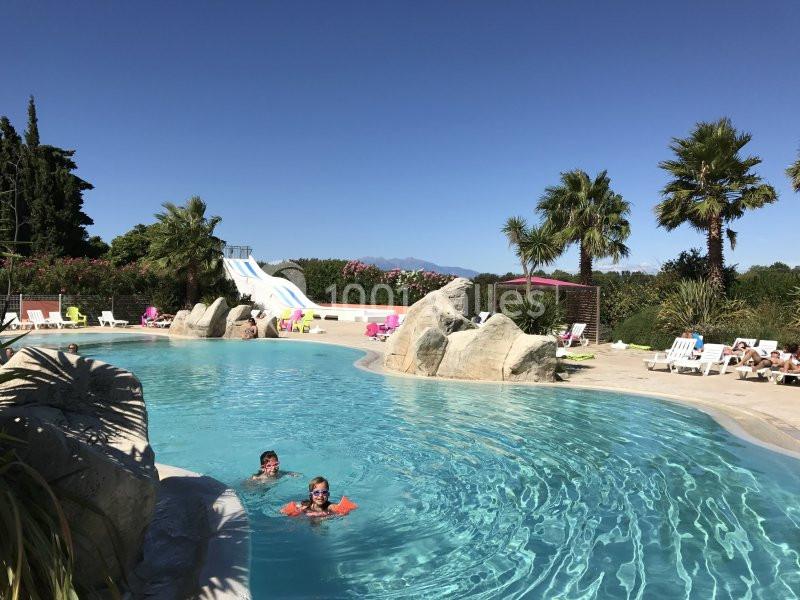 Piscine extérieure avec toboggans, palmiers et transats, deux enfants nageant sous un ciel dégagé.