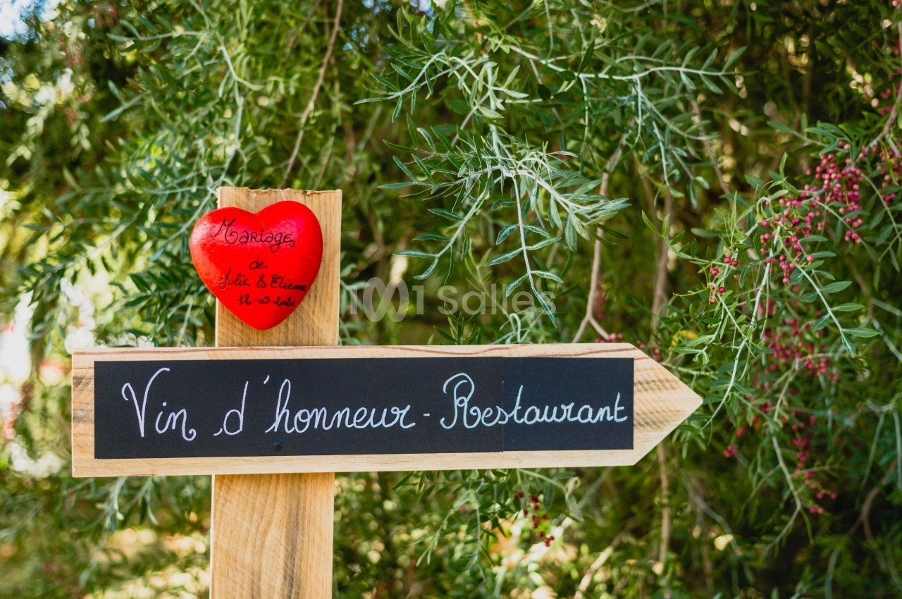 Panneau en bois indiquant ’Vin d'honneur - Restaurant’ avec un cœur rouge suspendu, devant un fond végétal.