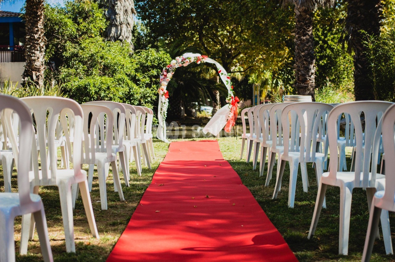 Allée centrale avec tapis rouge menant à une arche fleurie, entourée de chaises blanches en extérieur.