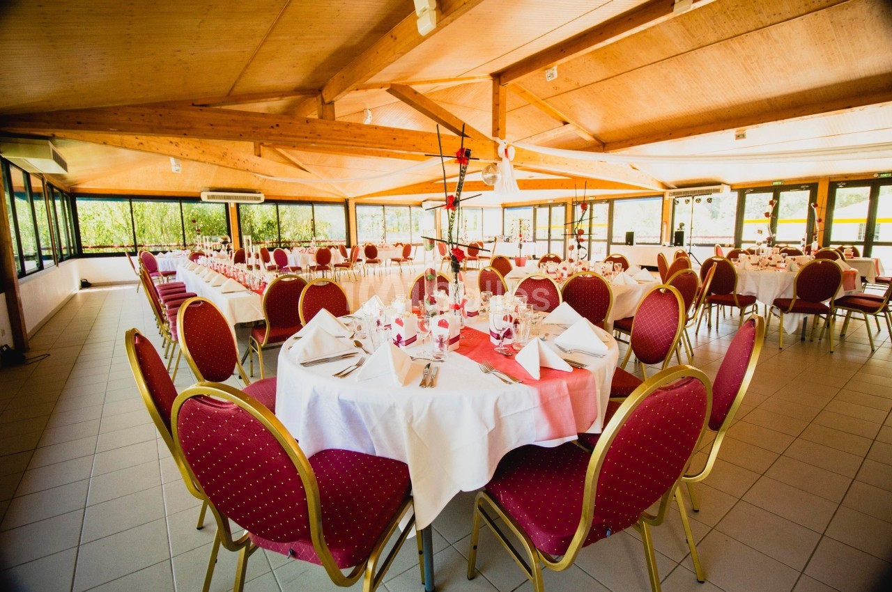 Salle de réception lumineuse avec tables rondes dressées, nappes blanches et chaises rouges, entourée de baies vitrées.