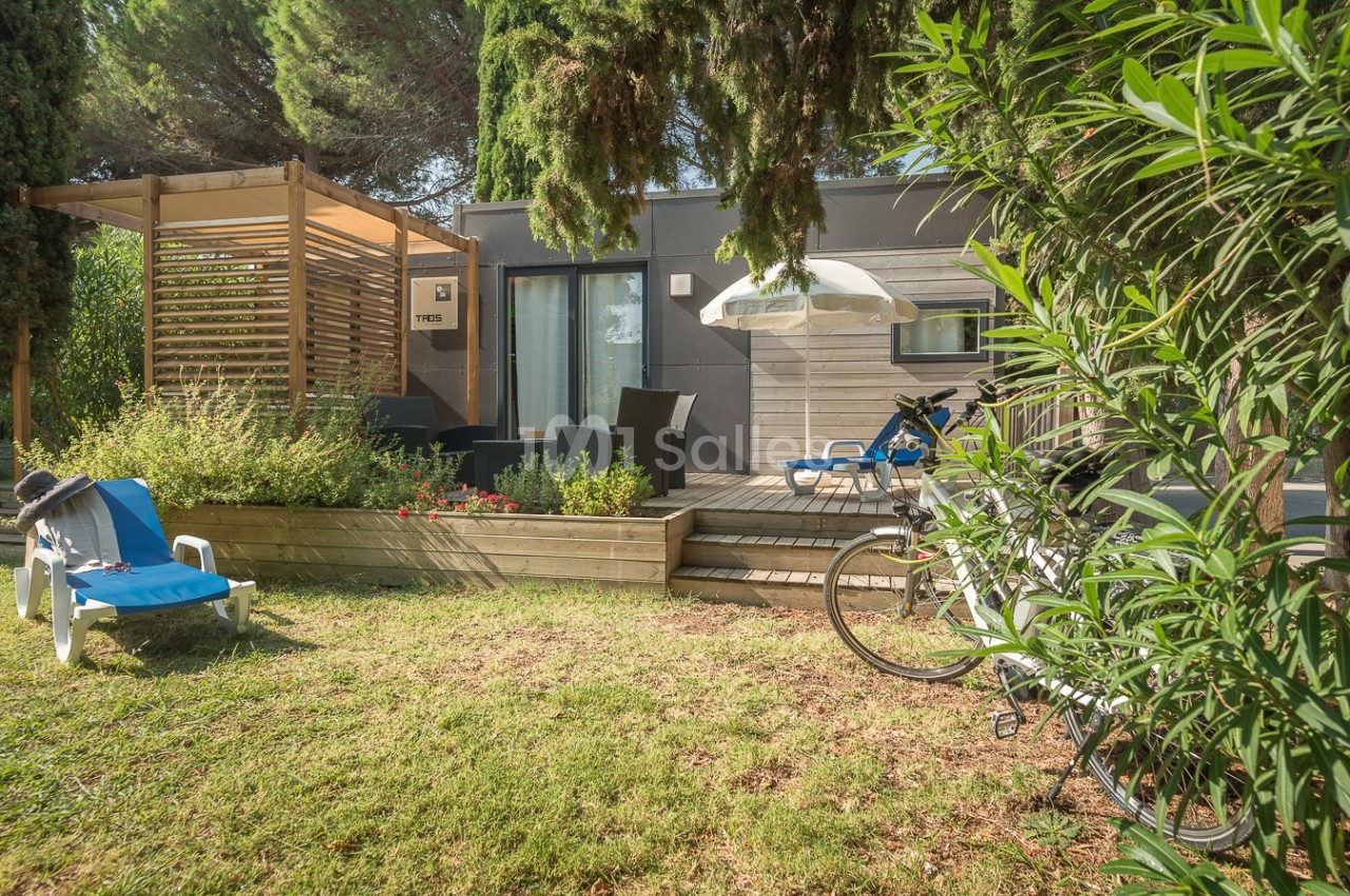 Espace extérieur avec terrasse en bois, chaises longues, vélos et végétation entourant une habitation moderne.