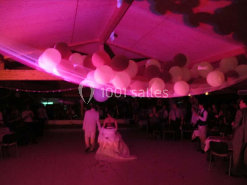 Un couple marche sous un plafond décoré de ballons roses et blancs, entouré d'invités dans une salle faiblement éclairée.