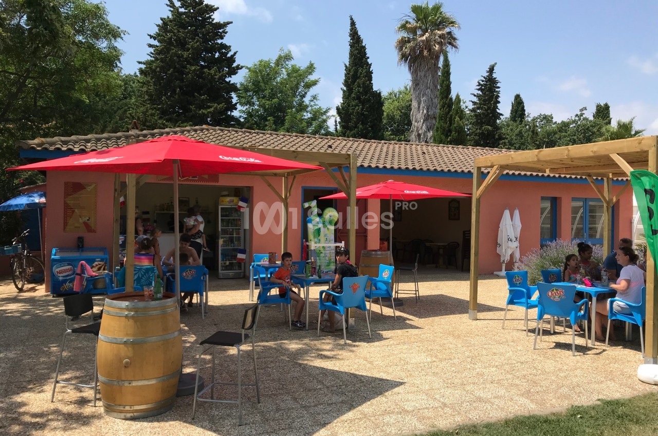 Terrasse d'un café avec des tables, chaises bleues et parasols rouges, entourée de végétation et d'arbres.