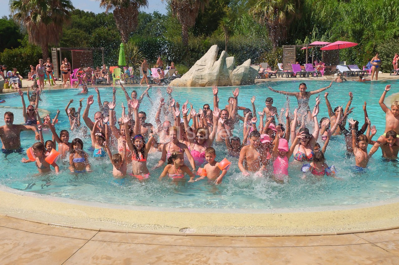 Groupe de personnes s'amusant et éclaboussant dans une piscine extérieure entourée de végétation et de parasols.